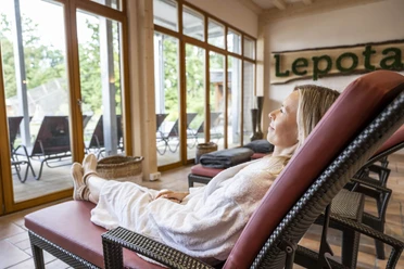 Loma_Juli2022_036.jpg Eine Frau entspannt in einem Spa-Bereich auf einer Liege, umgeben von großen Fenstern mit Gartenblick.
