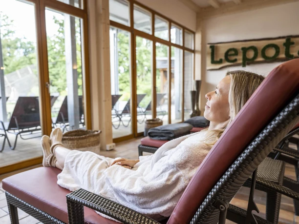 Loma_Juli2022_036.jpg Eine Frau entspannt in einem Spa-Bereich auf einer Liege, umgeben von großen Fenstern mit Gartenblick.