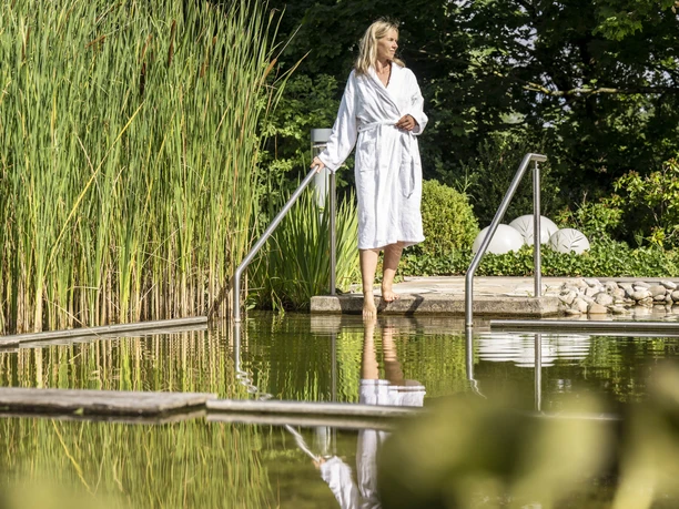 Loma_Juli2022_367.jpg Eine Frau im Bademantel geht an einem natürlichen Schwimmteich entlang, umgeben von Schilf.