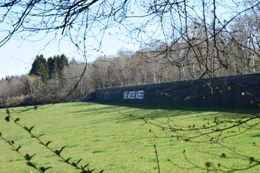Mauer am Aussichtspunkt "Die Mauer" Auf dem Bild ist eine Mauer auf einem Feld, mit dem Schriftzug "Nie wieder Krieg", vor Bäumen zu sehen.