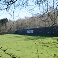 Mauer am Aussichtspunkt "Die Mauer" Auf dem Bild ist eine Mauer auf einem Feld, mit dem Schriftzug "Nie wieder Krieg", vor Bäumen zu sehen.