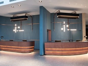 Hilton Cologne FO Moderne Hotelrezeption mit zwei geschwungenen, beleuchteten Empfangstresen und eleganter Beleuchtung.</p>Modern hotel reception with two curved, illuminated reception counters and elegant lighting.</p> <p