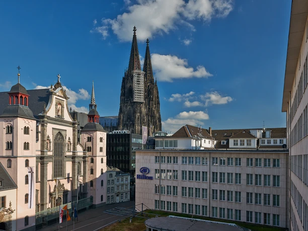 Hilton Cologne Kölner Dom mit steinernen Türmen und umliegenden Gebäuden bei blauem Himmel und leichter Bewölkung.