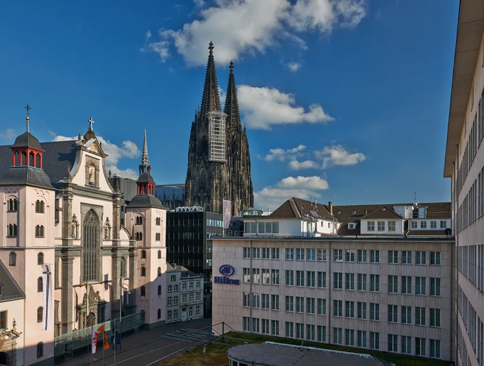 Hilton Cologne Kölner Dom mit steinernen Türmen und umliegenden Gebäuden bei blauem Himmel und leichter Bewölkung.Cologne Cathedral with stone towers and surrounding buildings under a blue sky and light cloud cover.