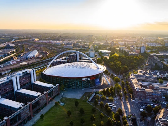 Bild Arena Luftaufnahme des LANXESS arena in Köln bei Sonnenuntergang, umgeben von städtischer Architektur.</p>Aerial view of the LANXESS arena in Cologne at sunset, surrounded by urban architecture.</p> <p