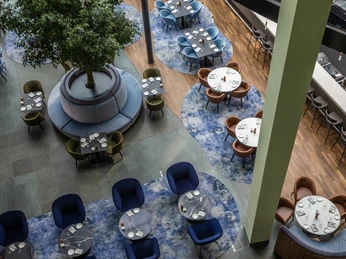 Bar Luftaufnahme einer modernen Hotelbar, ausgestattet mit runden Tischen und blauen Teppichen. Ein großer Baum im Zentrum sorgt für Atmosphäre, während bunte Stühle harmonisch arrangiert sind.Aerial view of a modern hotel bar, furnished with round tables and blue carpets. A large tree in the center creates atmosphere, while colorful chairs are harmoniously arranged.