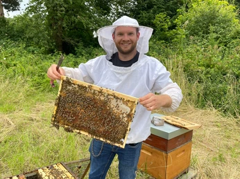 Die NeanderBiene in Aktion Mann im Imkeranzug hält eine Wabe voller Bienen; mit Bäumen und Wiese im Hintergrund.