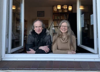 Fenstermusik mit Stefan Römer Stefan Römer und eine Frau blicken lächelnd durch ein geöffnetes Fenster mit Büchern im Hintergrund.