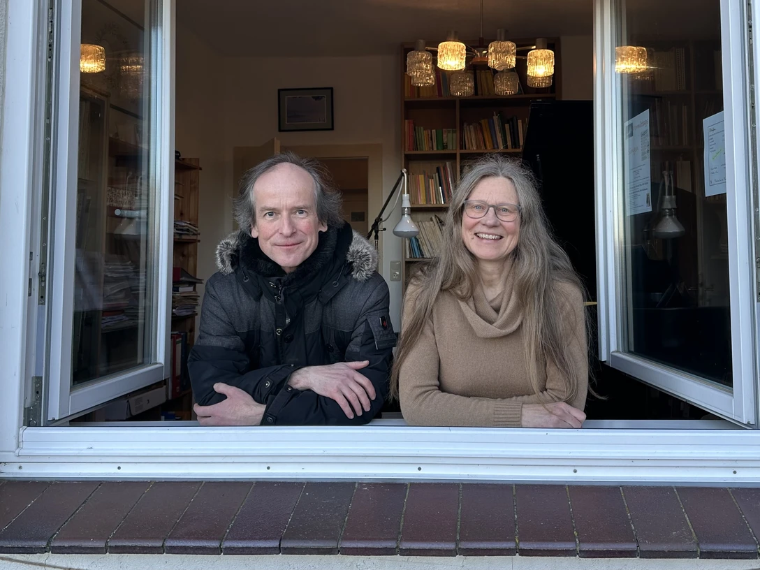 Fenstermusik mit Stefan Römer Stefan Römer und eine Frau blicken lächelnd durch ein geöffnetes Fenster mit Büchern im Hintergrund.