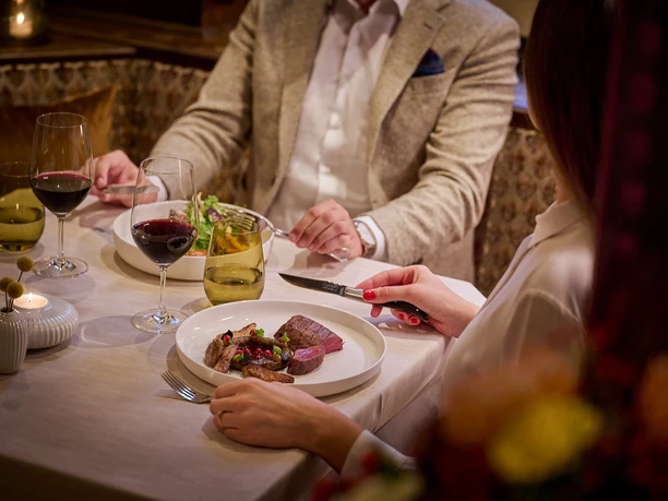Zwei elegant gekleidete Personen genießen ein stilvolles Abendessen mit Wein und Fleischgerichten.