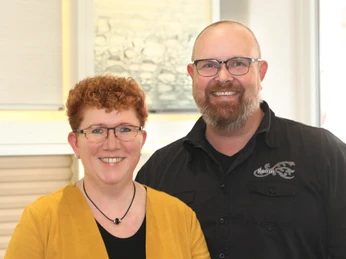 Raumausstattung Weist Ein lächelndes Paar, beide mit Brille. Die Frau trägt eine gelbe Jacke, der Mann ein schwarzes Hemd.