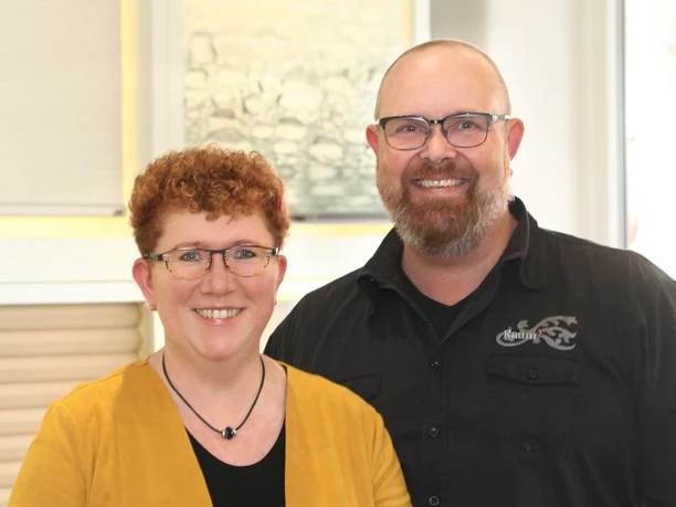 Raumausstattung Weist Ein lächelndes Paar, beide mit Brille. Die Frau trägt eine gelbe Jacke, der Mann ein schwarzes Hemd.