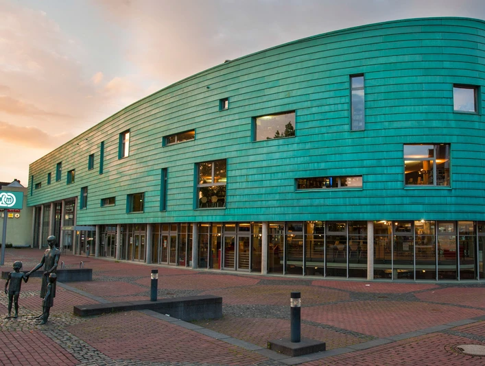MEDIO Aussenansicht Daemmerung Futuristisches Gebäude mit grüner Fassade und Glasfront bei Sonnenuntergang auf einem Platz.</p>Futuristic building with green façade and glass front at sunset on a square.</p> <p