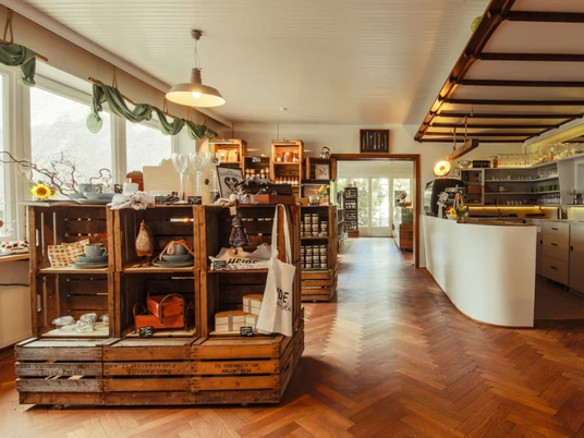 Degenhof_Manufaktur_.jpg Das Bild zeigt ein gemütliches Ladengeschäft mit rustikalem Interieur: Holzkisten gefüllt mit handgefertigten Waren, eine elegante, geschwungene Verkaufstheke und tiefe Holzfußböden, die eine warme Atmosphäre schaffen. Große Fenster lassen natürliches Licht einfallen, wodurch der Raum einladend und freundlich wirkt. Der Laden steht für traditionelle Handwerkskunst kombiniert mit modernem Design.
