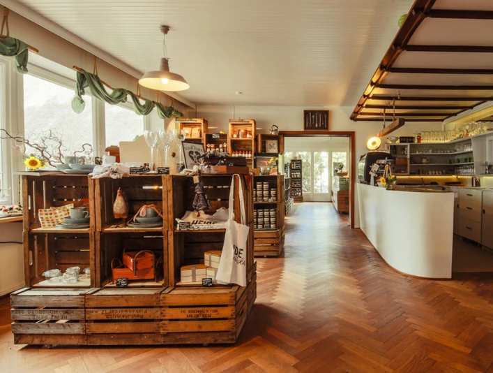 Degenhof_Manufaktur_.jpg Das Bild zeigt ein gemütliches Ladengeschäft mit rustikalem Interieur: Holzkisten gefüllt mit handgefertigten Waren, eine elegante, geschwungene Verkaufstheke und tiefe Holzfußböden, die eine warme Atmosphäre schaffen. Große Fenster lassen natürliches Licht einfallen, wodurch der Raum einladend und freundlich wirkt. Der Laden steht für traditionelle Handwerkskunst kombiniert mit modernem Design.