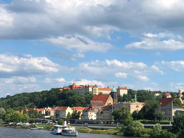 City view with river in the foreground, historic buildings with red roofs and green tower, surrounded by trees under a blue sky with clouds.
