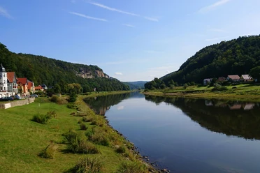 Blick vom Elbufer in Wehlen Richtung Rathen Flusslandschaft mit ruhigem Wasser, grünen Ufern und bewaldeten Hügeln; links bunte Häuser, klare, sonnige Atmosphäre.