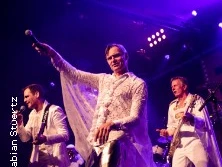 Eine Band in weißen Kostümen performt auf einer Bühne, der Sänger hält ein Mikrofon hoch.