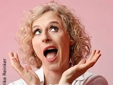 Eine Frau mit lockigem, blondem Haar zeigt eine freudige Überraschungsgeste vor rosa Hintergrund.