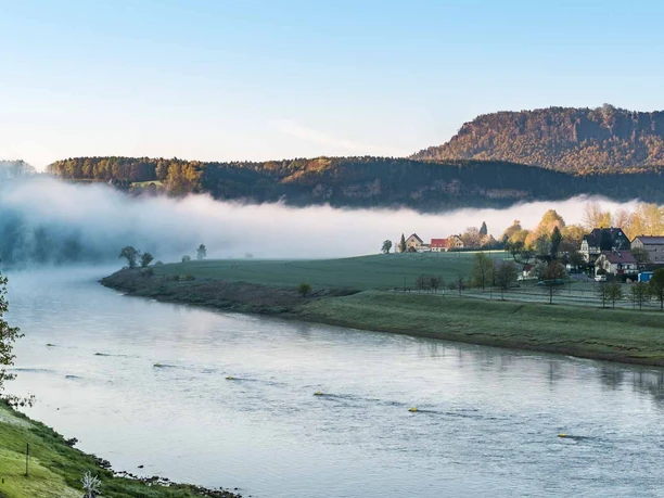 River landscape in the morning mist with a village on the bank, surrounded by green meadows and a wooded hill in the background under a clear sky.