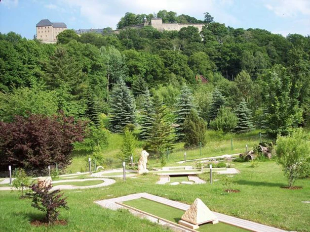 Minigolfing with a view of Königstein Fortress Minigolf course in a green landscape with trees and bushes, in the background a castle on a wooded hill.
