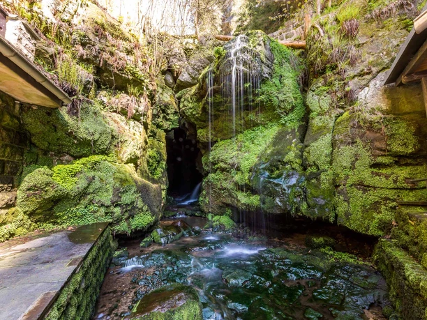 A small waterfall flows over moss-covered rocks into a cave surrounded by lush vegetation and stone walls, in a tranquil, natural setting.