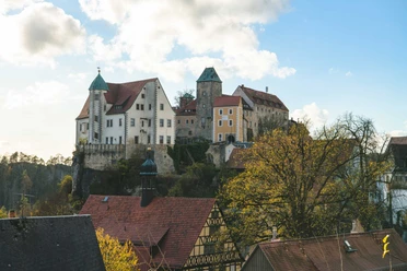 A historic castle with red roofs sits enthroned on a hill, surrounded by trees and traditional half-timbered houses under a blue sky.