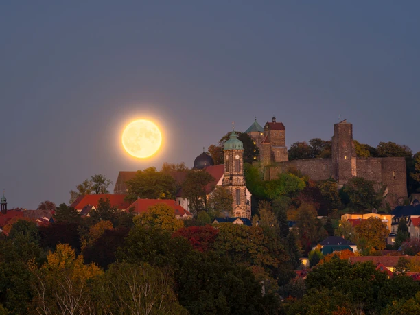 Volle maan boven Kasteel Stolpen met torens en rode daken, omringd door herfstbomen; rustige, sfeervolle schemering.