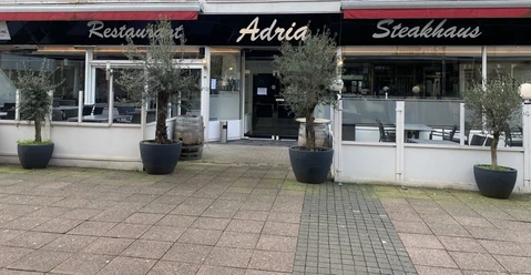 Außenansicht eines Restaurants mit der Aufschrift „Adria“. Drei Olivenbäume stehen vor dem Eingang.