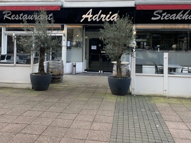 Außenansicht eines Restaurants mit der Aufschrift „Adria“. Drei Olivenbäume stehen vor dem Eingang.