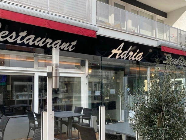 Steakhaus & Restaurant Adria mit moderner Glasfassade und Außenbestuhlung unter roten Markisen.