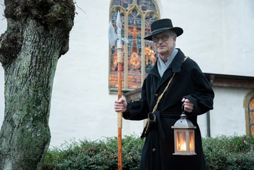 Ein Nachtwächter in traditionellem Gewand mit Laterne und Hellebarde vor einer historischen Kirche.