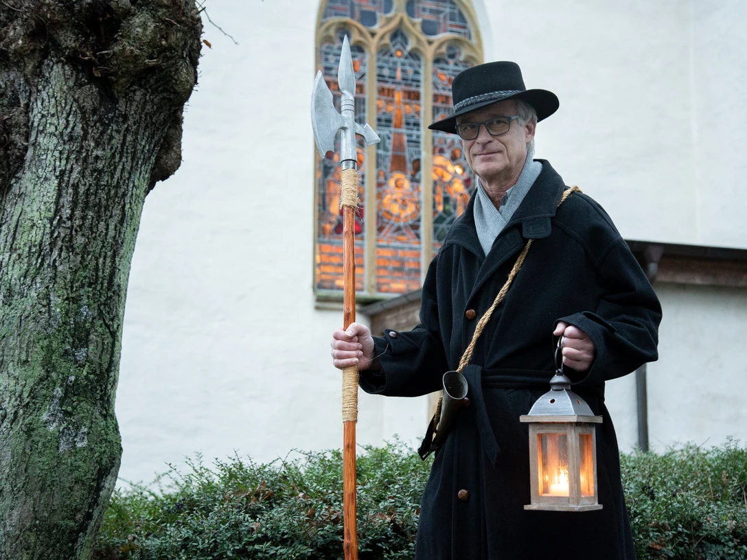 Nachtwächter Roland Zöller (1).jpg Ein Nachtwächter in traditionellem Gewand mit Laterne und Hellebarde vor einer historischen Kirche.