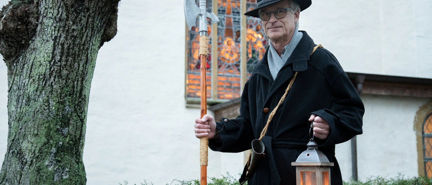 Nachtwächter Roland Zöller (1).jpg Ein Nachtwächter in traditionellem Gewand mit Laterne und Hellebarde vor einer historischen Kirche.
