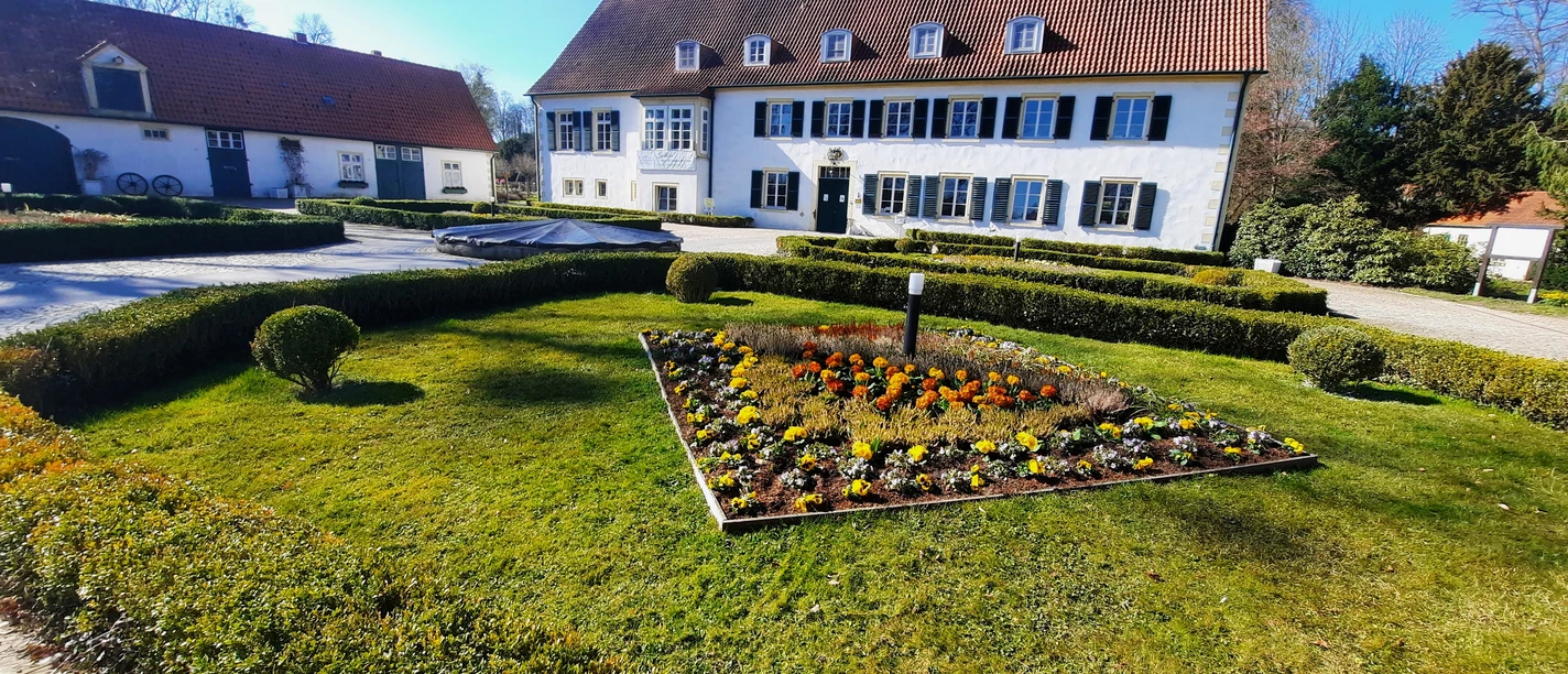 Haus des Gastes FrühjahrTouristik Preußisch Oldendorf.jpg Ein weißes Gebäude mit rotem Ziegeldach und blauen Fensterläden umgeben von gepflegten Gärten.