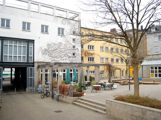 Ansicht Bürgerhaus Kalk aus dem hausinternen Hof Ein ruhiger städtischer Innenhof mit mehreren Gebäuden, Tischen und Stühlen im Freien.