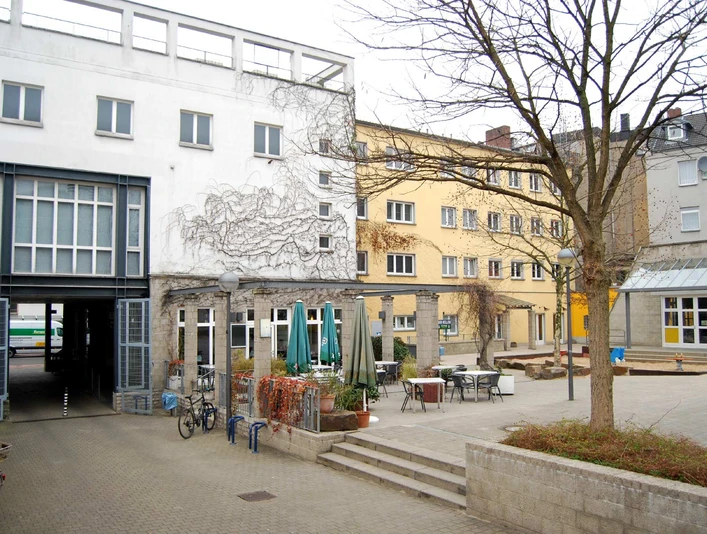 Ansicht Bürgerhaus Kalk aus dem hausinternen Hof Ein ruhiger städtischer Innenhof mit mehreren Gebäuden, Tischen und Stühlen im Freien.A quiet urban courtyard with several buildings, outdoor tables and chairs.
