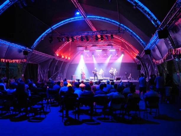 Open Air Bühne im Hof des BHK Open-Air-Bühne im Innenhof des BHK: Eine nächtliche Szene mit beleuchteter Bühne unter einem geschwungenen Zelt, umgeben von sitzendem Publikum.