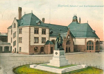 Historisches Bahnhofsgebäude mit Uhrenturm und Denkmal in Herford, umgeben von gepflegtem Vorplatz.