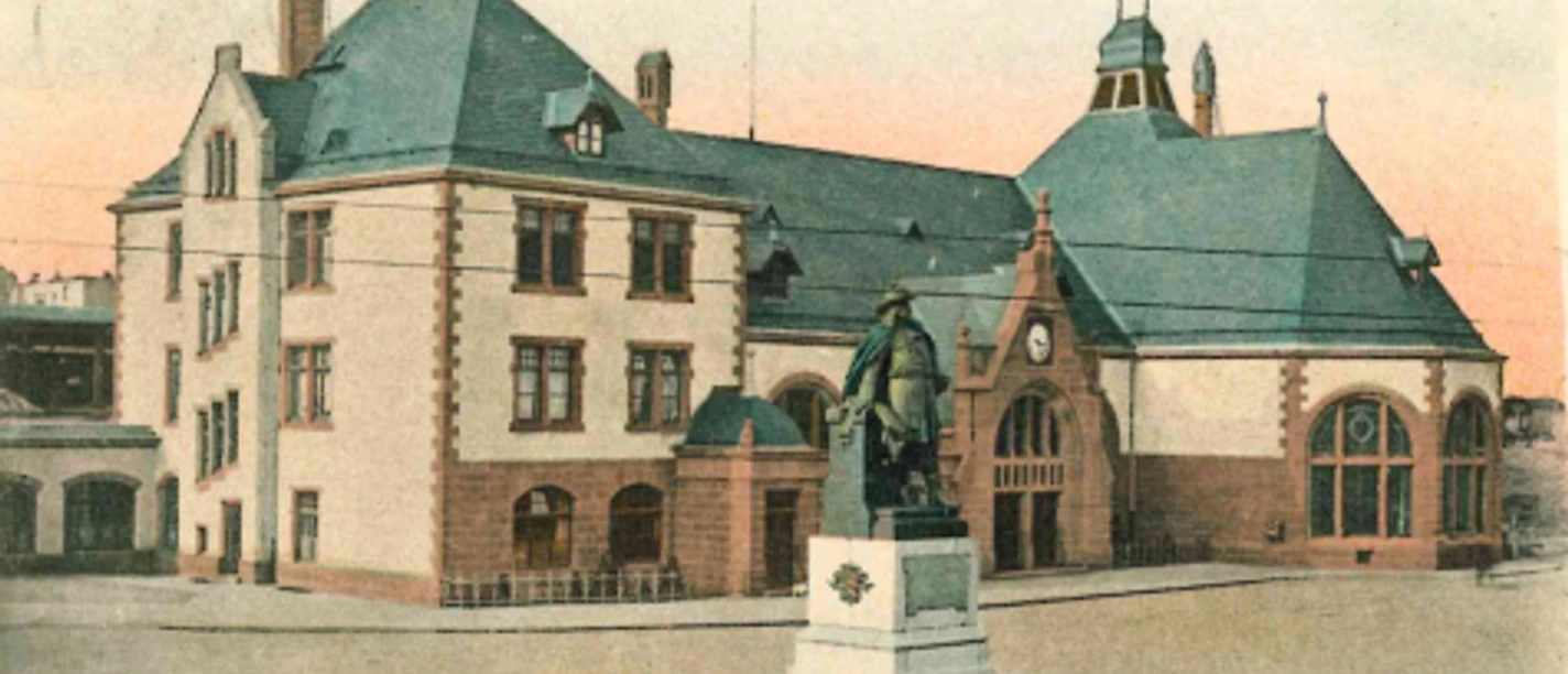 Historisches Bahnhofsgebäude mit Uhrenturm und Denkmal in Herford, umgeben von gepflegtem Vorplatz.
