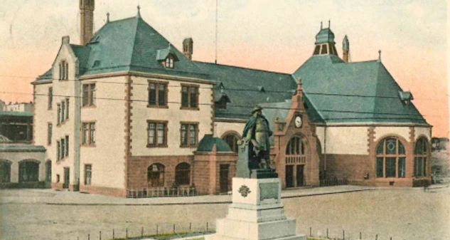 Historisches Bahnhofsgebäude mit Uhrenturm und Denkmal in Herford, umgeben von gepflegtem Vorplatz.