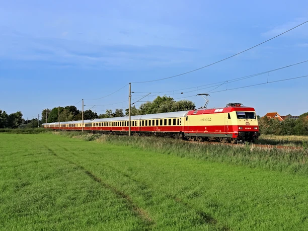 der private 1. Klasse-Sonderzug AKE-RHEINGOLD Schnellzug mit roter Lok fährt durch grüne Wiese bei klarem Himmel und Stromleitungen.