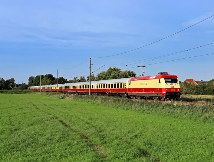 der private 1. Klasse-Sonderzug AKE-RHEINGOLD Schnellzug mit roter Lok fährt durch grüne Wiese bei klarem Himmel und Stromleitungen.Express train with red locomotive runs through green meadow with clear sky and power lines.
