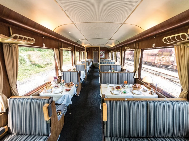 Speisewagen des AKE-RHEINGOLD Innenraum eines nostalgischen Speisewagens mit gedeckten Tischen, gestreiften Sitzen und Lampen.</p>