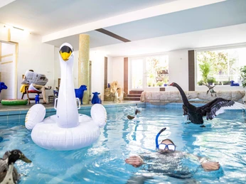 Indoor-Pool im Gartenhotel Fettehenne Ein lichtdurchfluteter Indoor-Pool mit Wasserfontäne. Ein Schwimmer trägt eine Taucherbrille. Ein aufblasbarer Schwan schwimmt im Pool, dekoriert mit Tierfiguren.