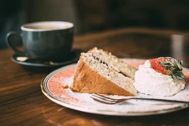 A slice of cake with cream and strawberries on a plate, a cup of coffee next to it on a wooden table.