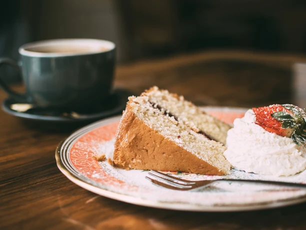 A slice of cake with cream and strawberries on a plate, a cup of coffee next to it on a wooden table.