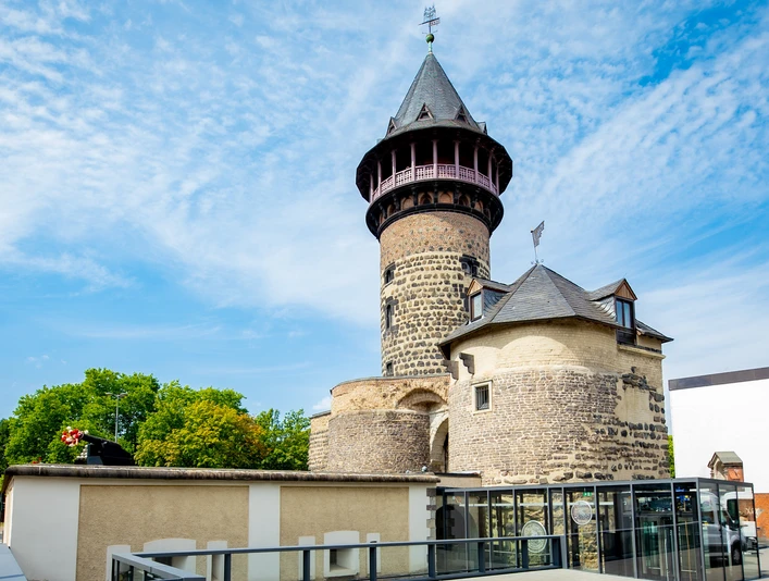 Eventlocation Ulrepforte Aussenansicht Alter Wasserturm in Mönchengladbach mit zylindrischem Steinbau und spitzem Dach vor blauem Himmel.Old water tower in Mönchengladbach with cylindrical stone structure and pointed roof against a blue sky.