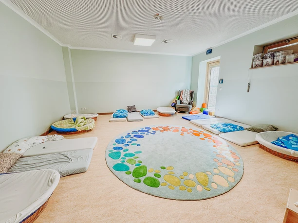 IMG_2024.jpg Heller Schlafraum im Kindergarten mit rundem, buntem Teppich und bodennahen Schlafplätzen.
