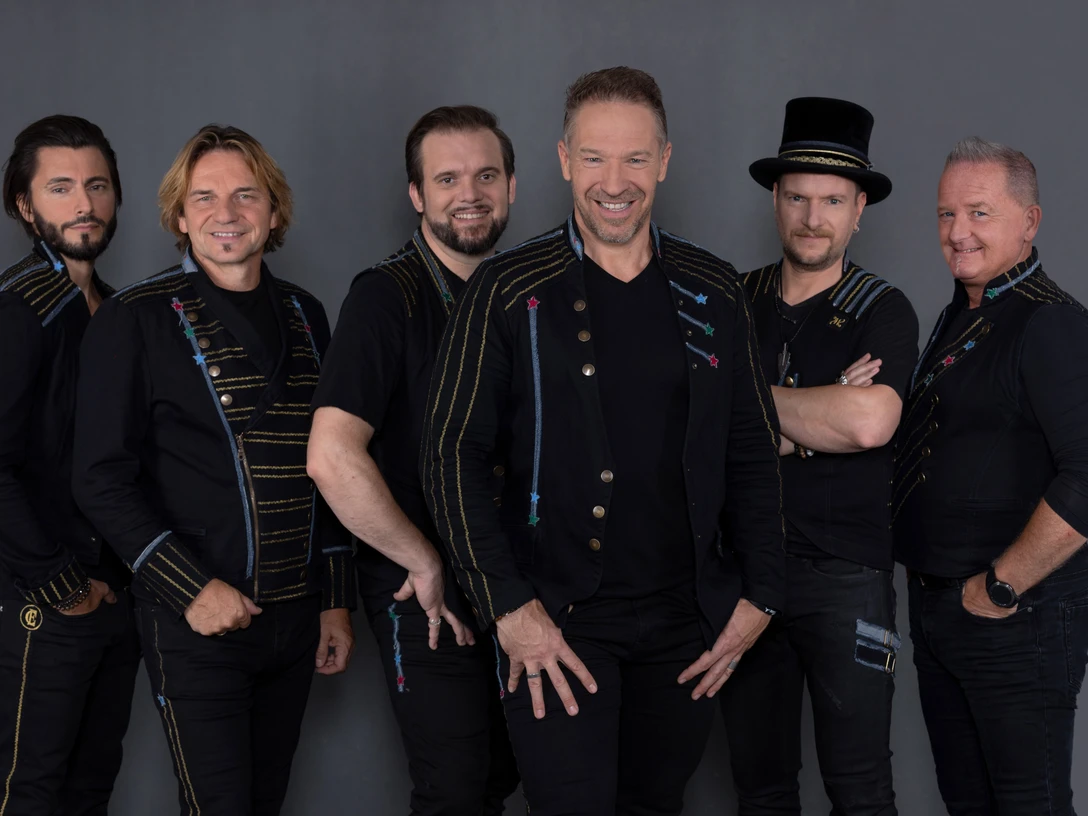 Höhner Sechs lächelnde Männer in schwarzer Kleidung und festlichem Design posieren vor grauem Hintergrund.</p>Six smiling men in black clothing and festive designs pose in front of a gray background.</p> <p